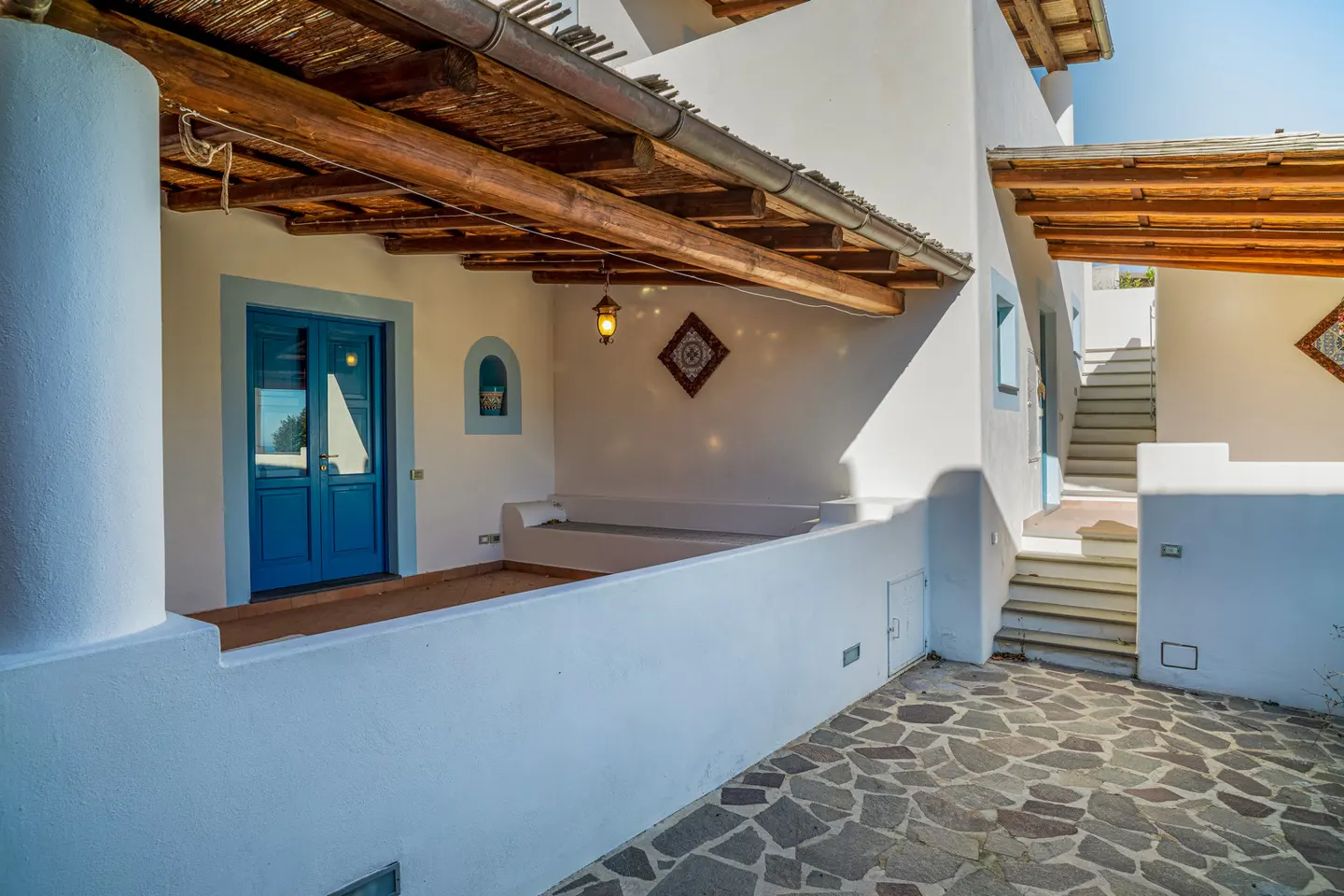 Exterior of a white building with a blue door and window frames, a wooden roof, and a stone-paved courtyard.
