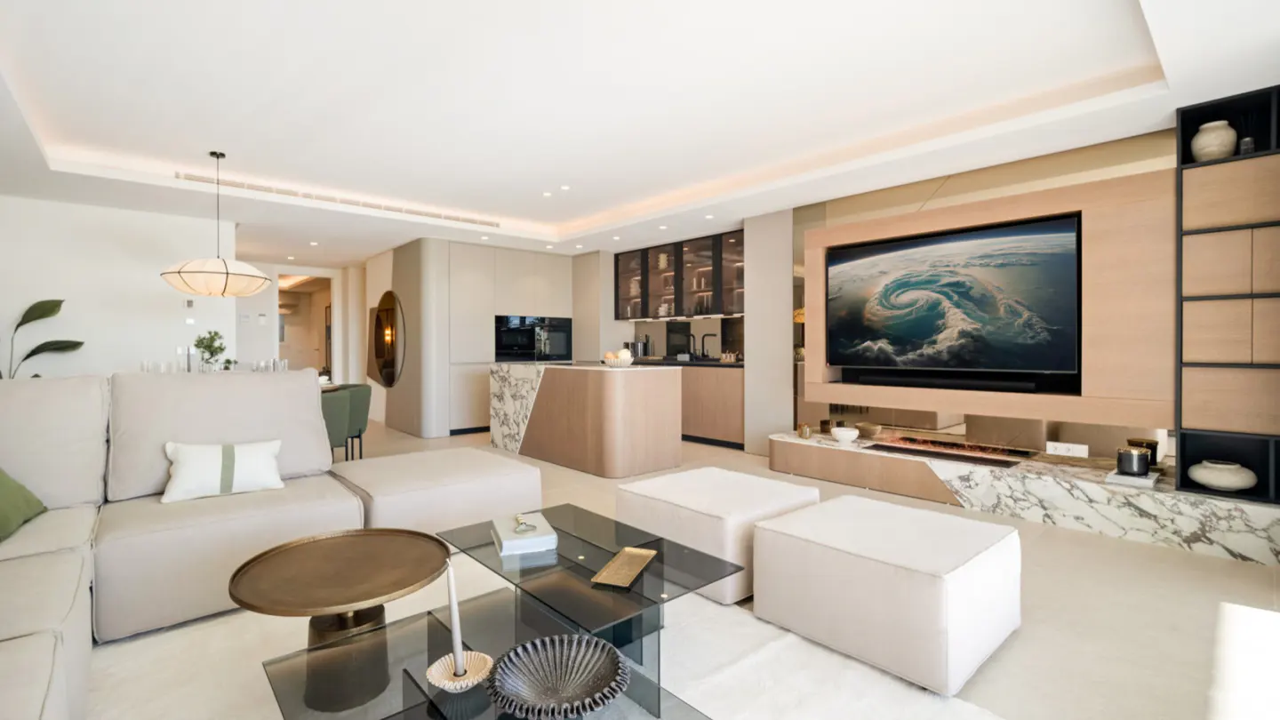 Bright, modern living room with beige sectional sofa, glass coffee table, and large TV showing a swirling storm. Kitchen area visible in the background.