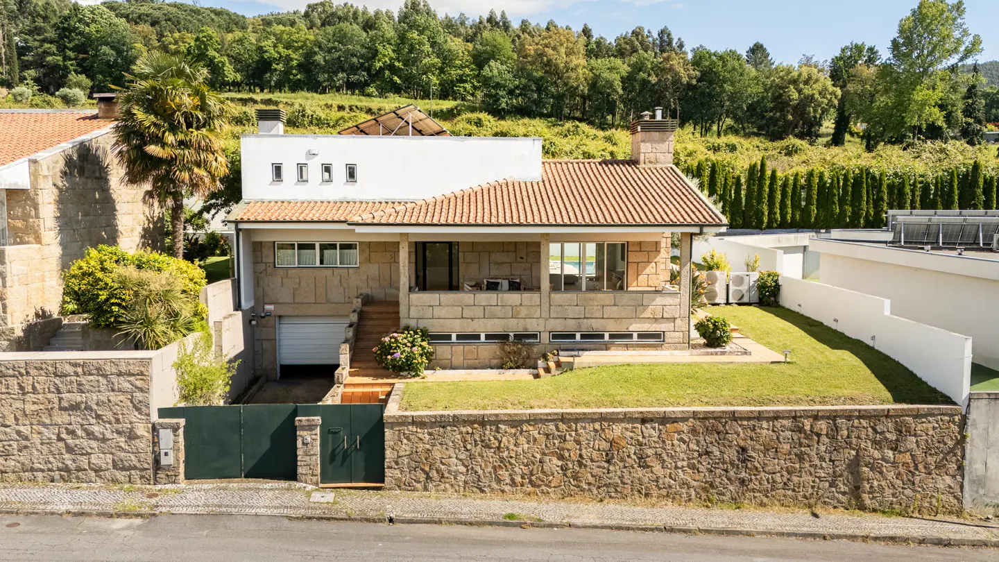 A two-story stone house with a terracotta roof and green gate, set against a backdrop of lush green trees.