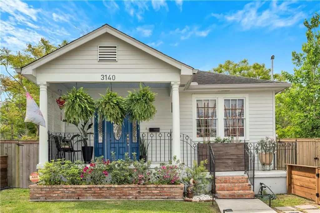 Exterior view of a white, single-story house with blue doors, hanging ferns, and a brick flower bed.