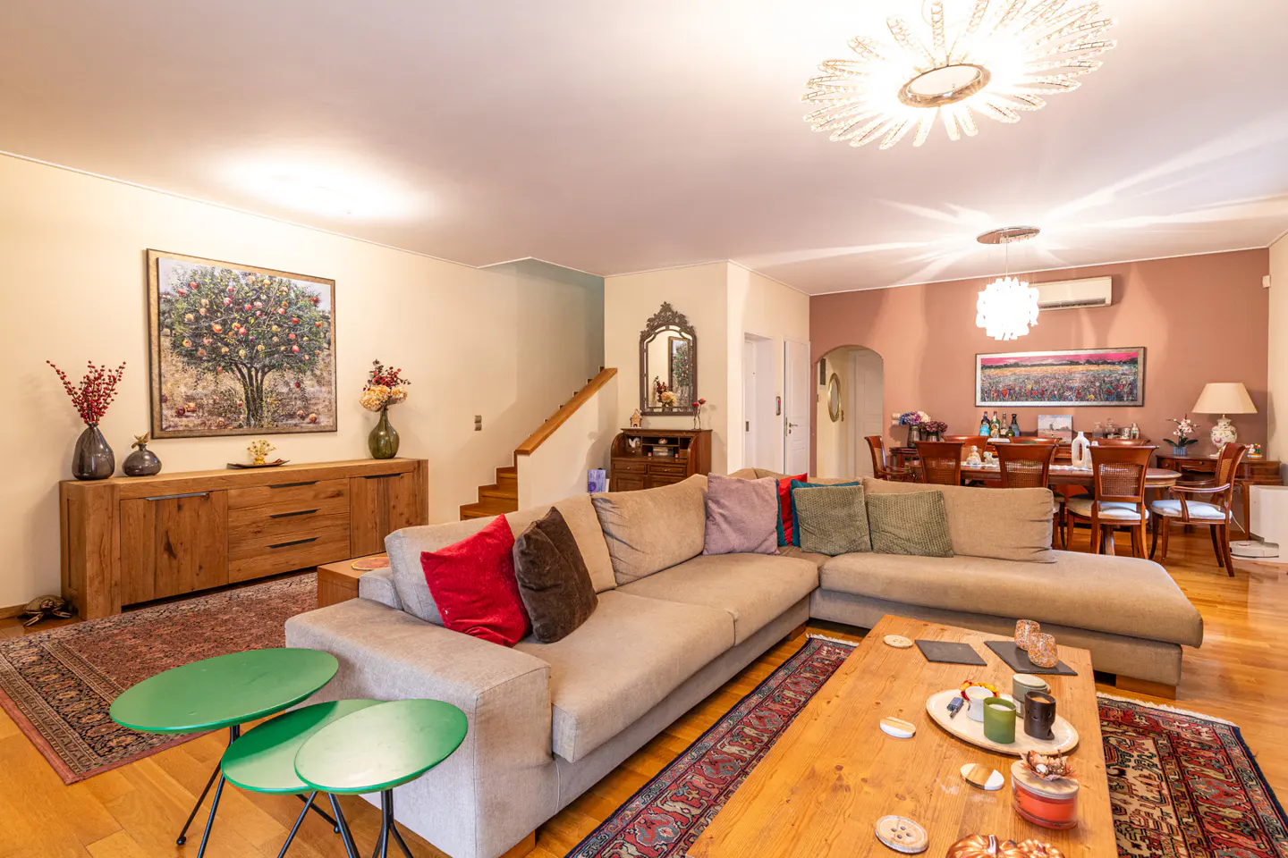 Bright living room with a beige sectional sofa, wooden floors, and a dining area in the background. Artwork and decorative items add to the homey feel.
