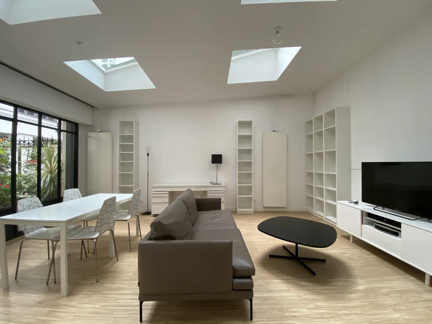 Bright, modern living room with skylights, white walls, and light wood floors. Features a gray sofa, black coffee table, and white dining set.