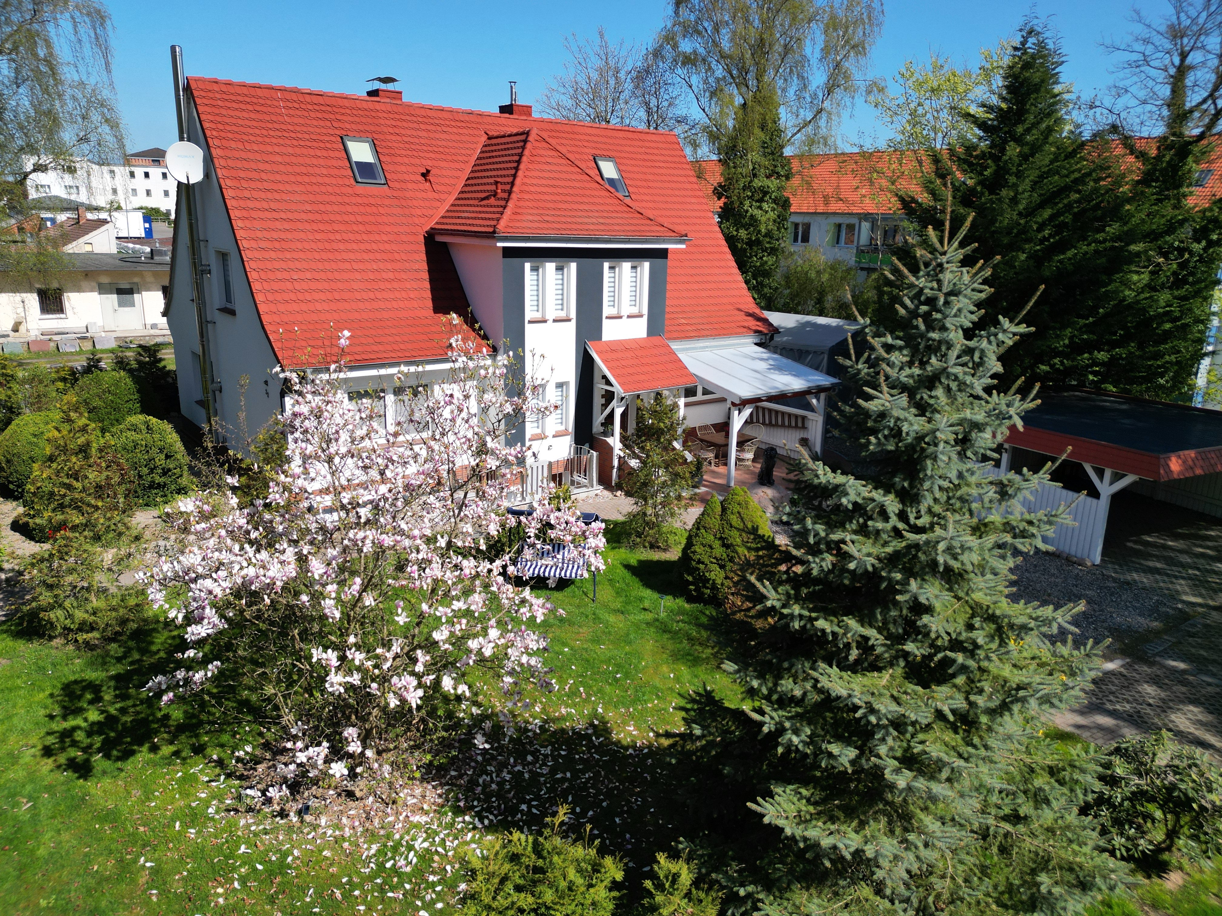 Exterior view of a two-story house with a red roof, white walls, and a green lawn with trees and flowers.