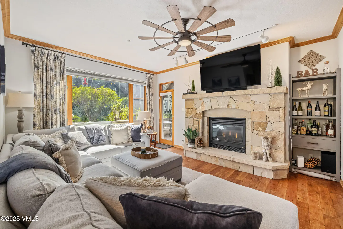 Living room with a large gray sectional sofa, stone fireplace with TV, and a bar area with liquor bottles.