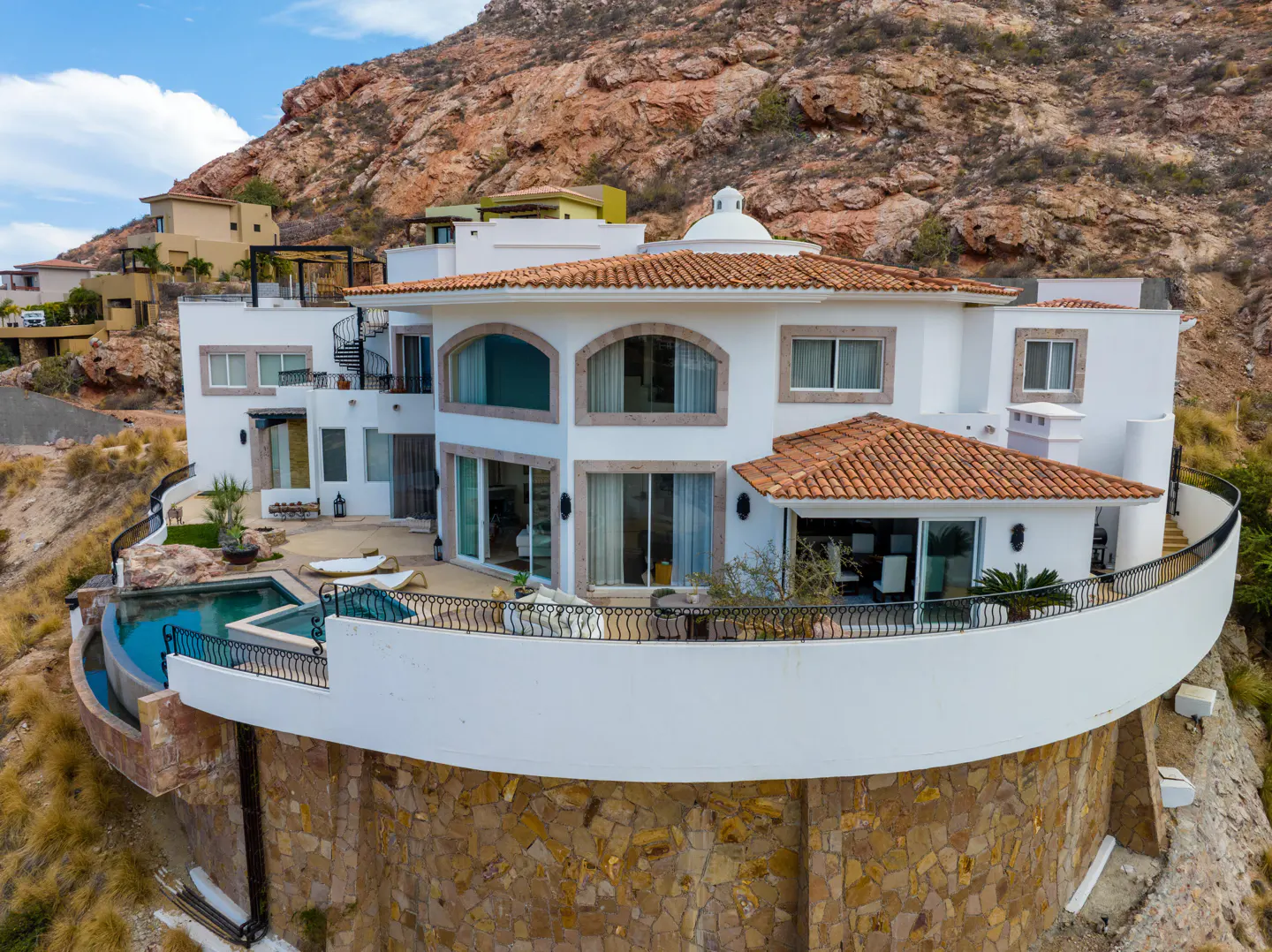 A white, multi-story house with a red tile roof sits on a hillside with a pool and patio.