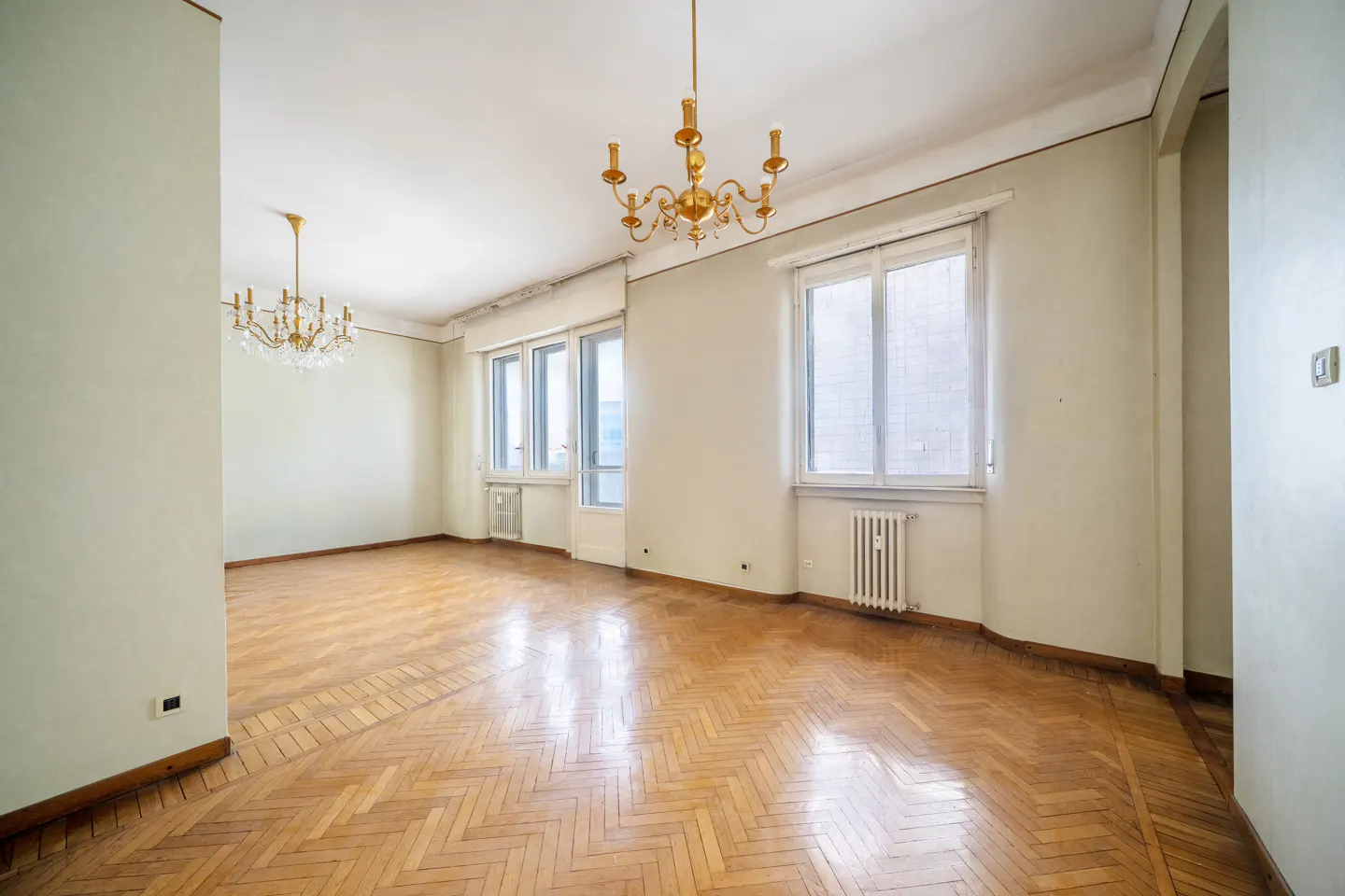 Empty room with herringbone wood floors, light green walls, and two gold chandeliers. Windows let in natural light.
