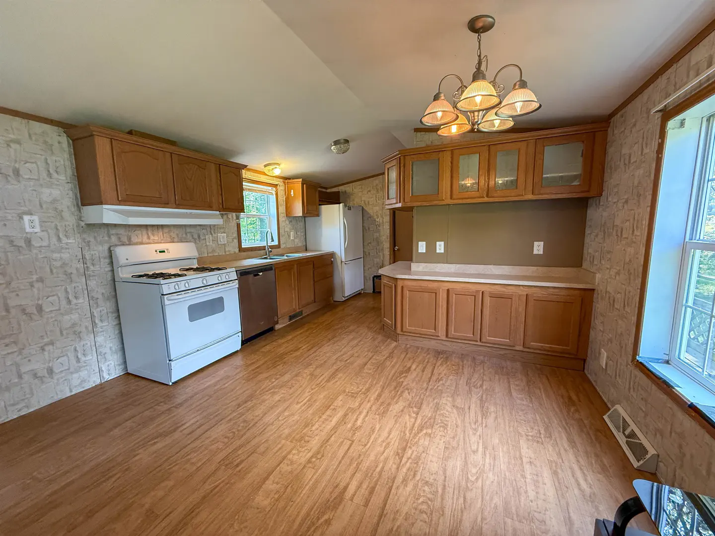 A bright kitchen with wood-look floors, brown cabinets, and a white stove. A chandelier hangs above a counter with cabinets.
