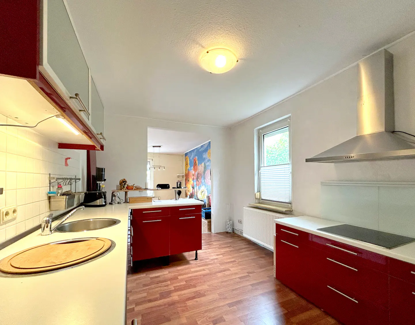 A bright kitchen with red cabinets, white countertops, and wood floors. A window and stainless steel range hood are visible.
