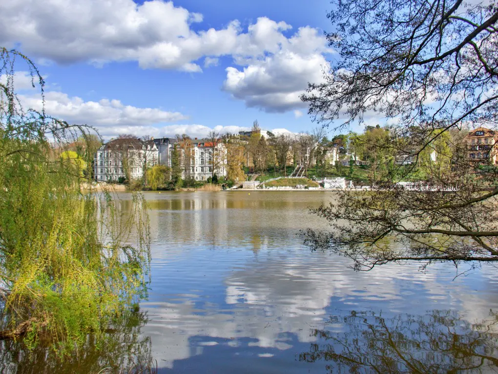 Scenic view of a lake reflecting blue sky and clouds, with buildings and trees in the background.