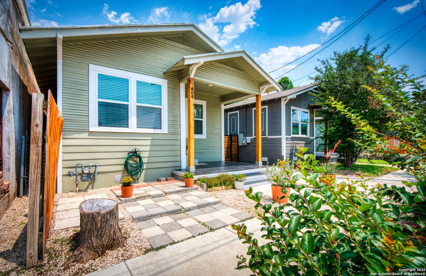 Exterior view of a light green house with a covered porch, stone walkway, and lush green landscaping under a blue sky.