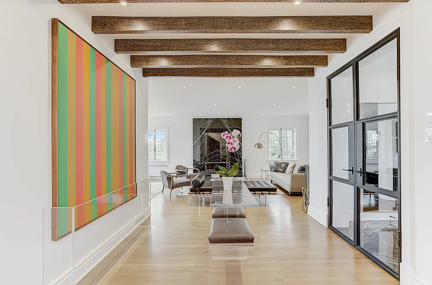 Bright foyer with wood floors, exposed beams, and a large colorful striped painting. A glass table and black framed glass doors are visible.