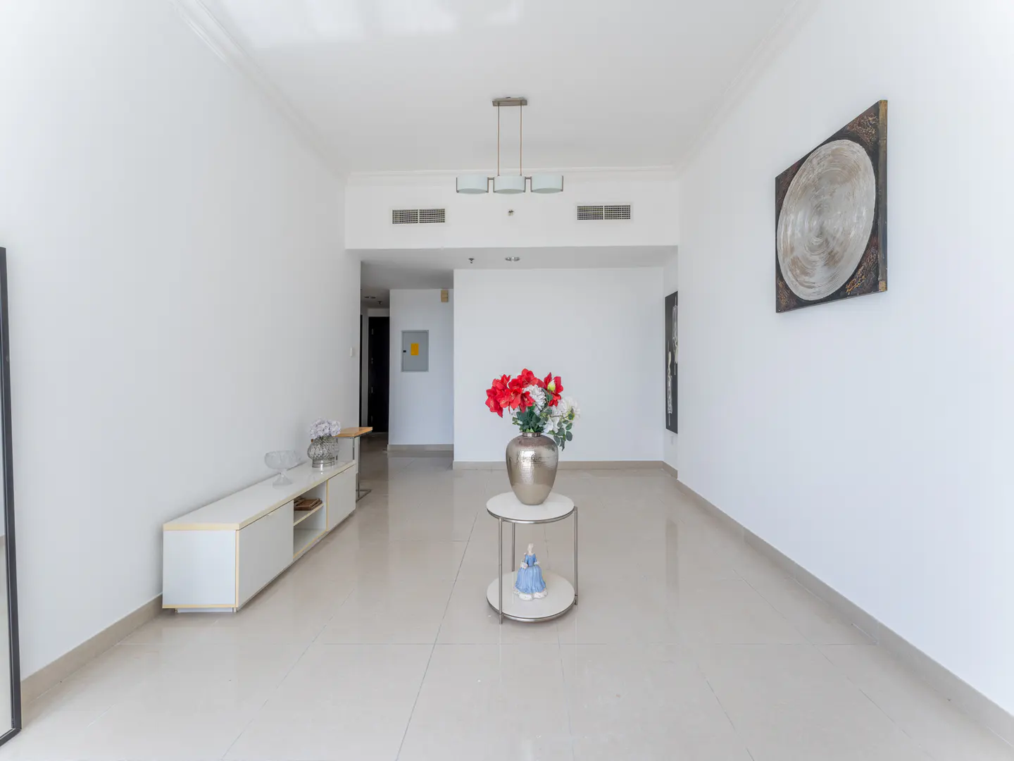 Bright, empty living room with white walls and tile floor. A silver vase with red flowers sits on a round table. A white media console is on the left.