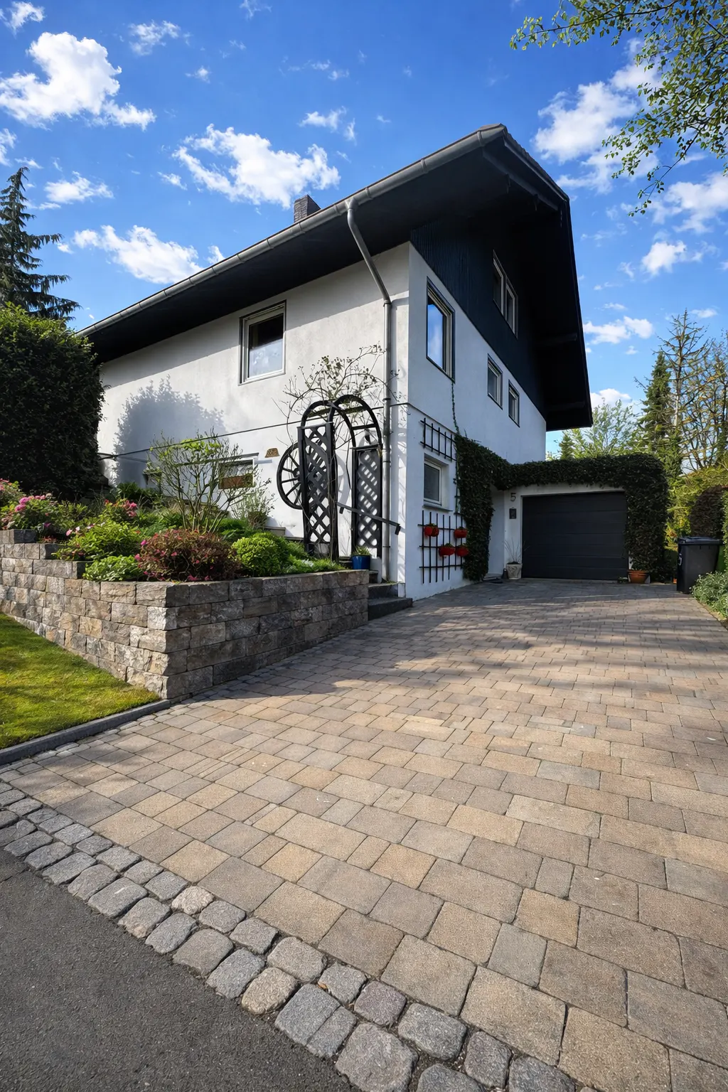 White house with dark roof, stone retaining wall, paved driveway, and garden arch under a bright blue sky.
