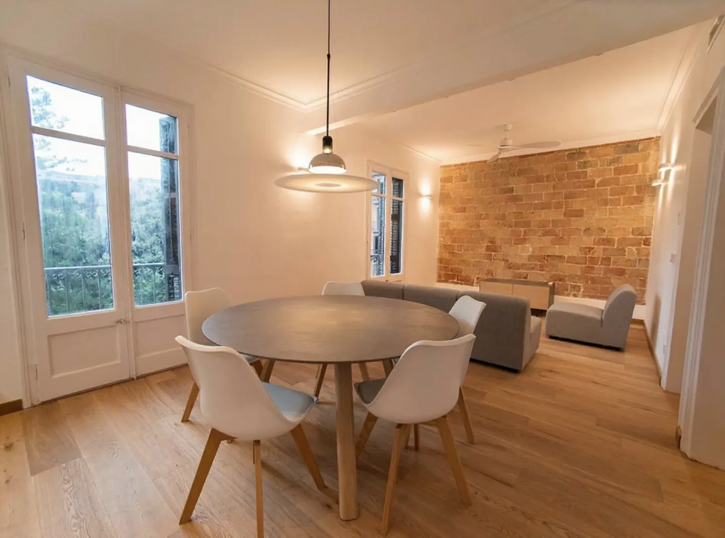 Bright living room with a round table, white chairs, and a brick wall. A gray sofa and light wood floors add to the modern design.