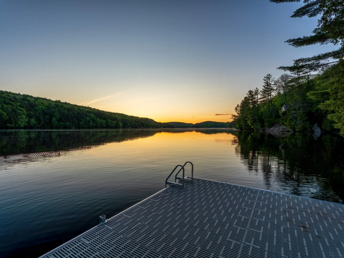 A gray floating dock with a ladder extends into a calm lake at sunset, surrounded by lush green forest-covered hills.