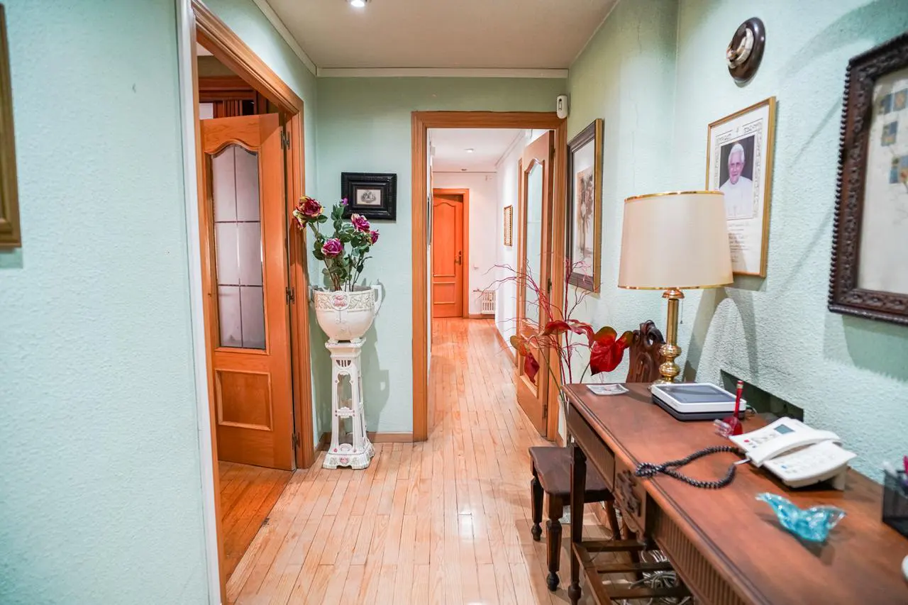 Hallway with light green walls, wood floors, and doors. A desk with a lamp and phone sits against the wall, adorned with framed pictures. A vase of flowers stands near an open door.