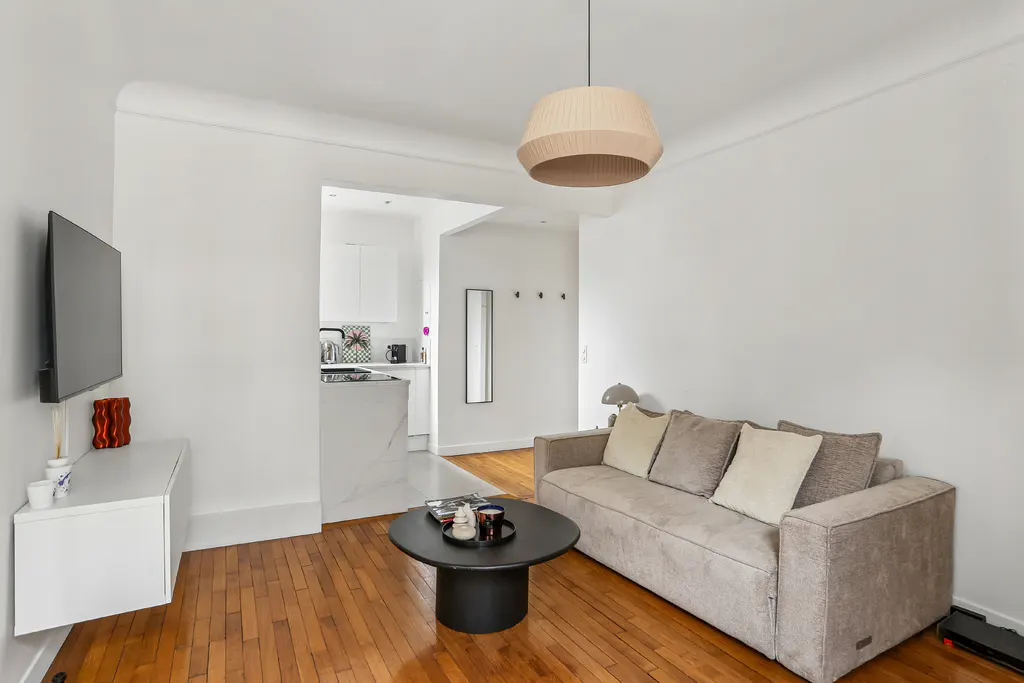 Modern living room with hardwood floors, a beige sofa, a round black coffee table, and a view into a white kitchen.