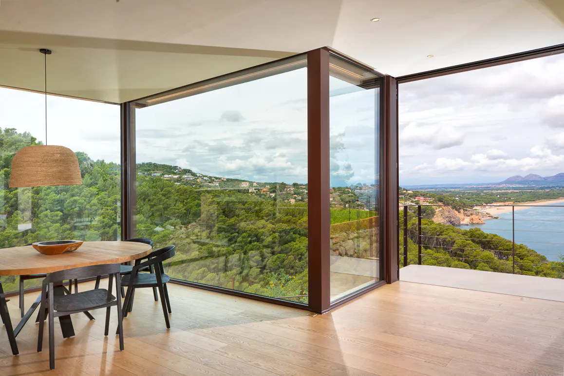 Modern dining area with wooden table and black chairs, featuring floor-to-ceiling windows overlooking a coastal forest.