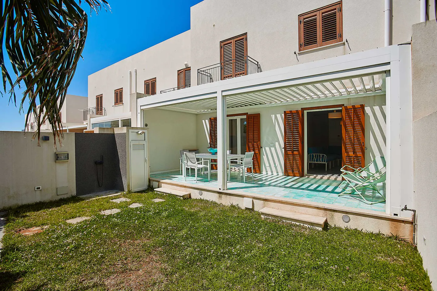 A sunny patio with a white pergola, dining set, and lounge chairs, overlooking a small grassy yard against a beige house.