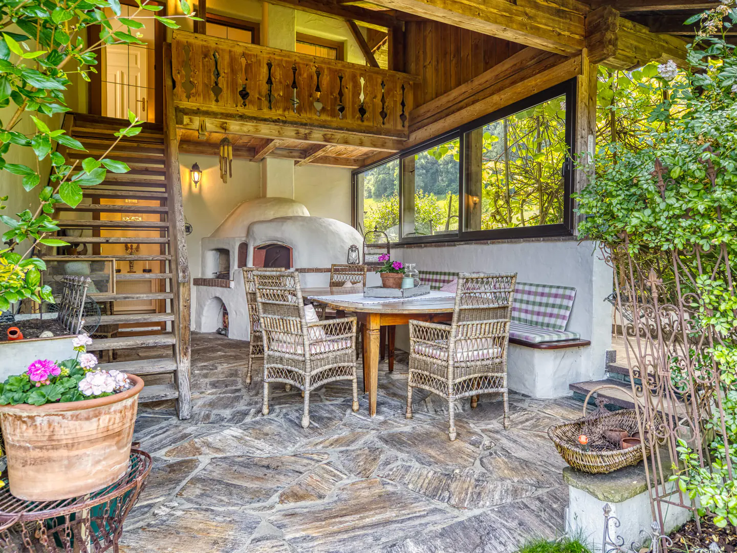 Outdoor patio with stone floor, wood table and chairs, pizza oven, and stairs to a balcony. Large window with green trees in background.