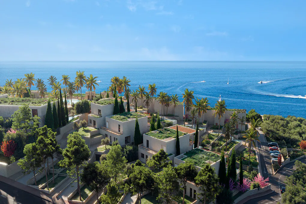 Aerial view of modern, light-colored villas with green roofs, palm trees, and ocean backdrop under a clear blue sky.