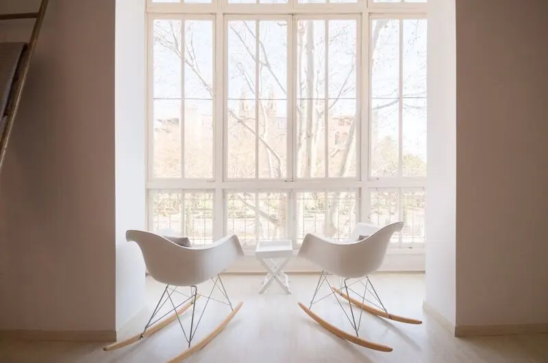 Bright room with two white rocking chairs facing a large window. A small white table sits between them. Outside, trees are visible.