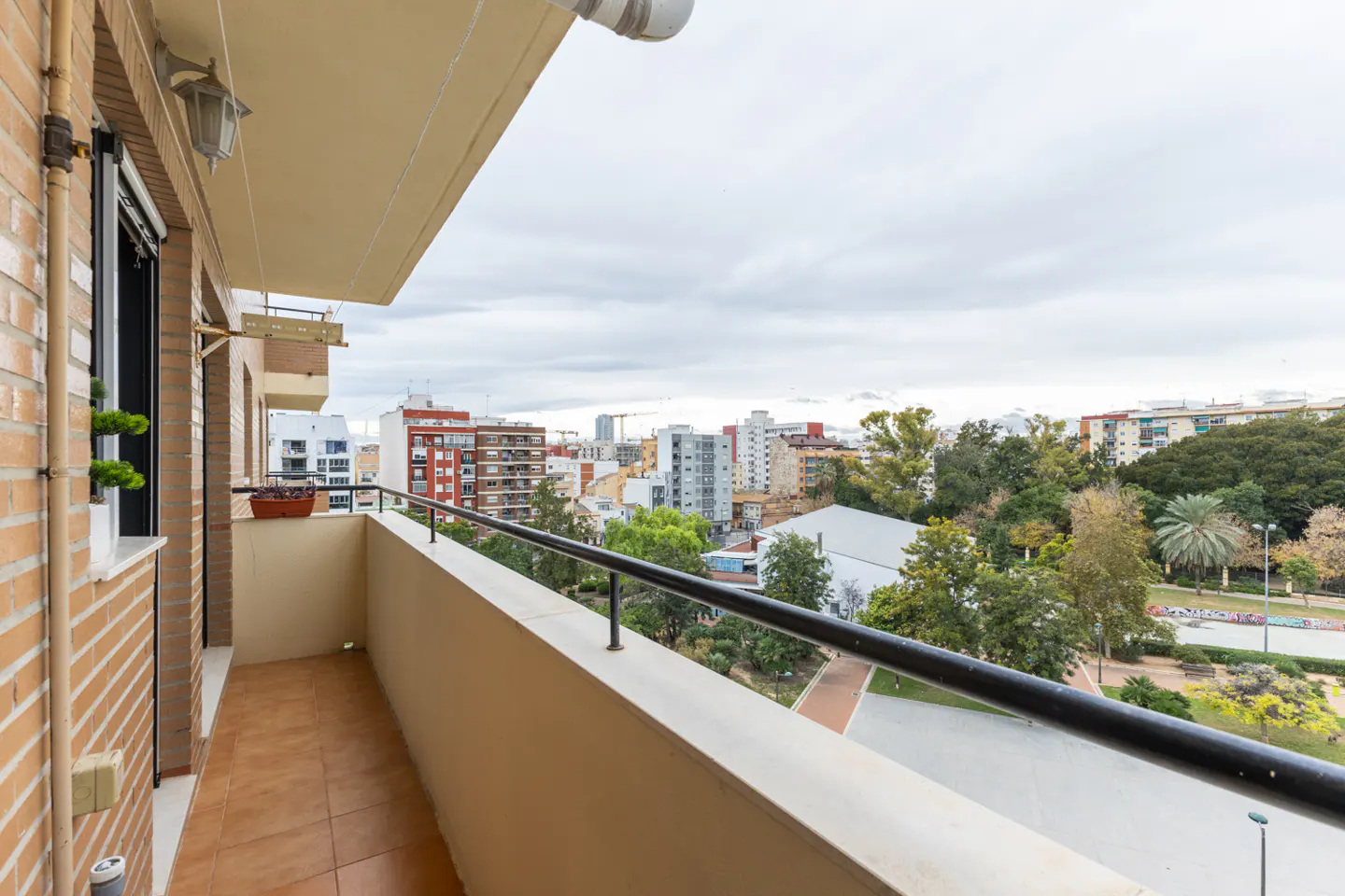 Balcony view of a city with buildings, trees, and a park under a cloudy sky. The balcony has a brown tile floor and a black metal railing.