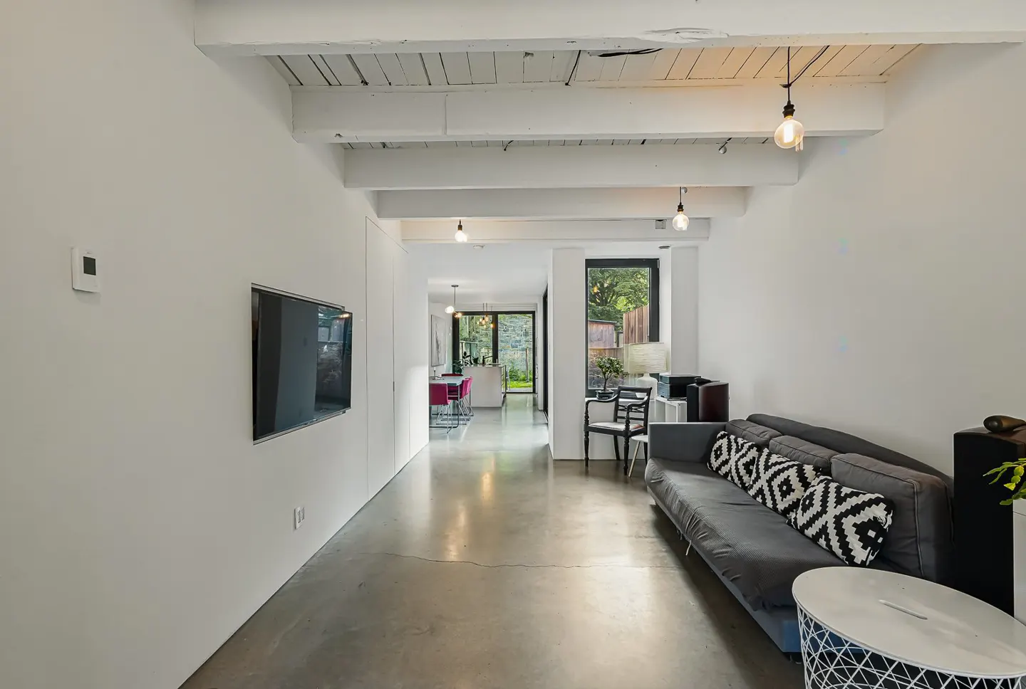 Bright, modern living room with white walls, concrete floors, and a gray sofa with patterned pillows. A TV is mounted on the wall.