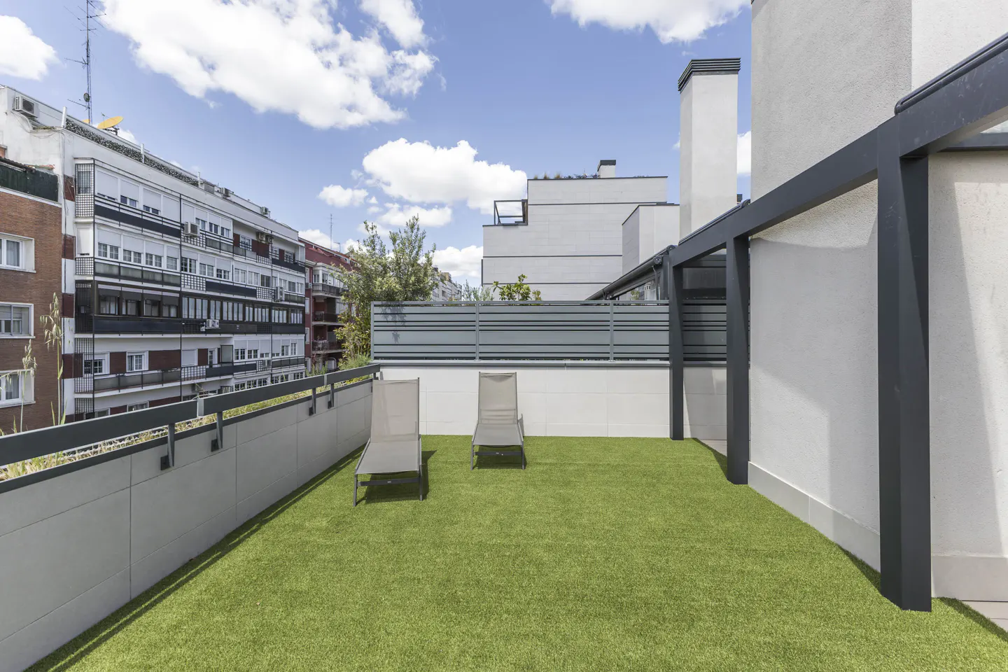 Rooftop terrace with artificial grass, two lounge chairs, and a dark pergola. Buildings and blue sky with clouds in the background.