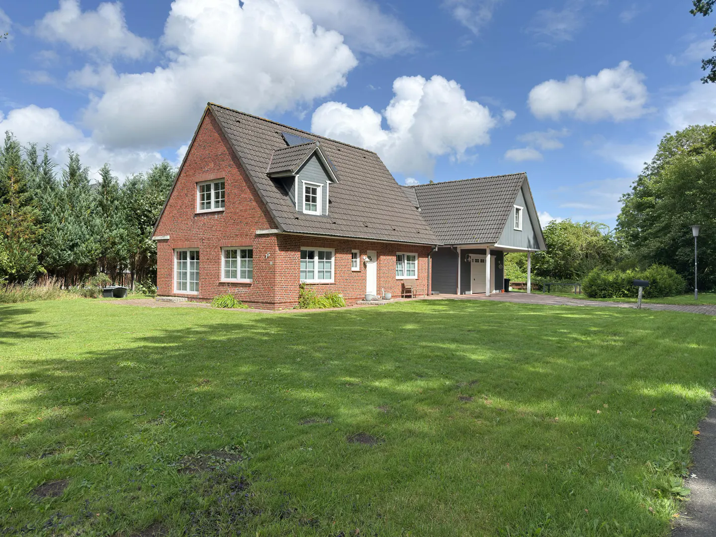 A two-story red brick house with a gray roof and white trim sits on a large green lawn under a blue sky with white clouds.