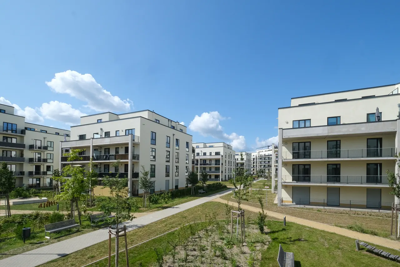 Modern apartment complex with beige buildings, balconies, green lawns, and paved walkways under a blue sky.