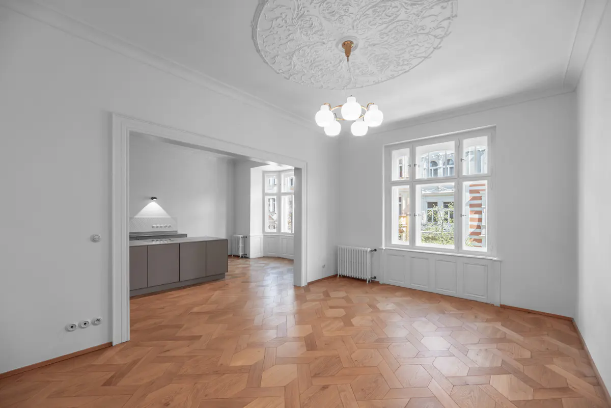 Bright, empty room with herringbone wood floors, white walls, and a modern chandelier. A kitchen is visible through an open doorway. Large windows let in natural light.