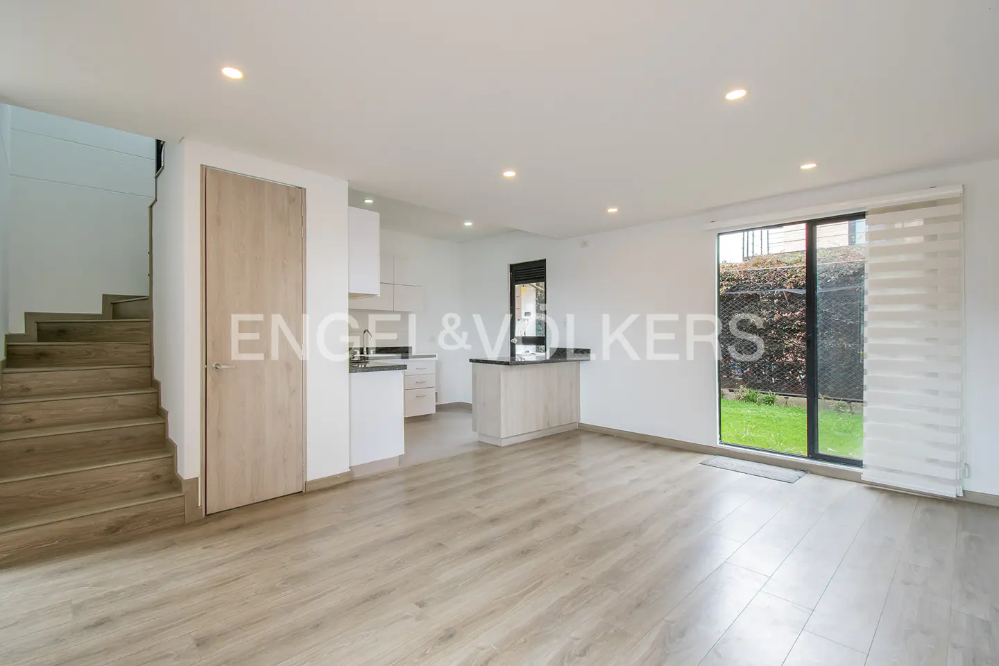 Bright, open-plan living space with wood-look floors, white walls, and a modern kitchen. Stairs lead to the upper level. Sliding glass doors open to a green lawn.