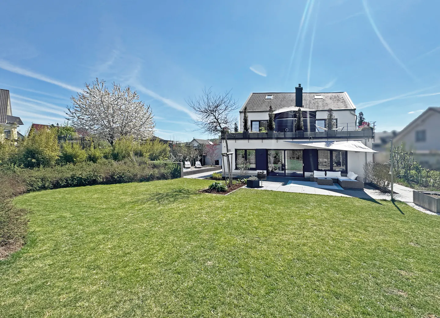 A modern two-story house with a green lawn, patio furniture, and a blooming white tree under a blue sky.
