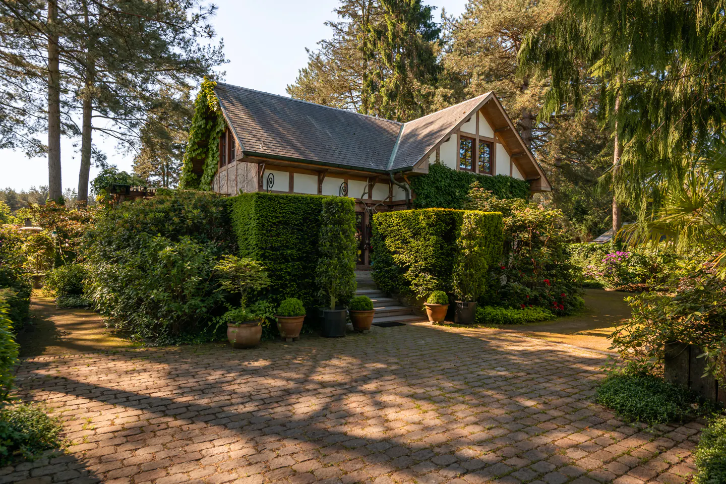 A charming half-timbered cottage with ivy-covered walls, surrounded by lush gardens and tall trees on a sunny day.
