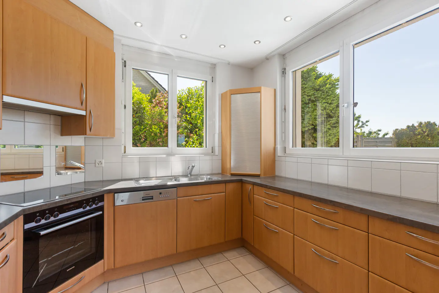 Modern kitchen with light wood cabinets, dark countertops, white tiled backsplash, built-in oven, and bright windows.