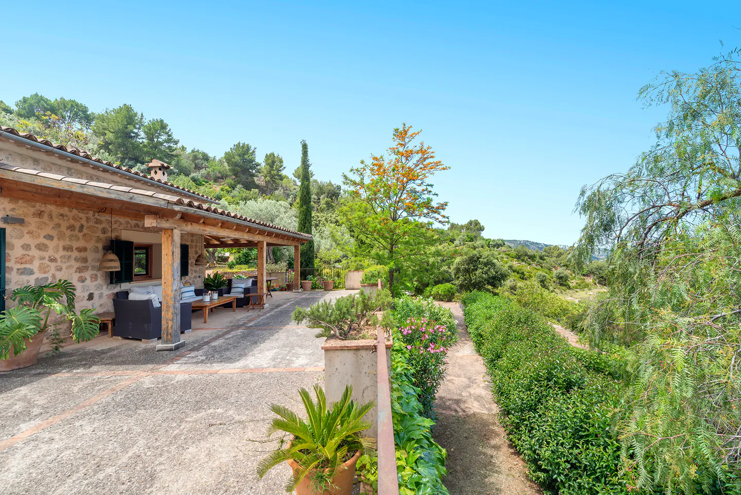 Exterior view of a stone house with a covered patio, surrounded by lush greenery and a clear blue sky.