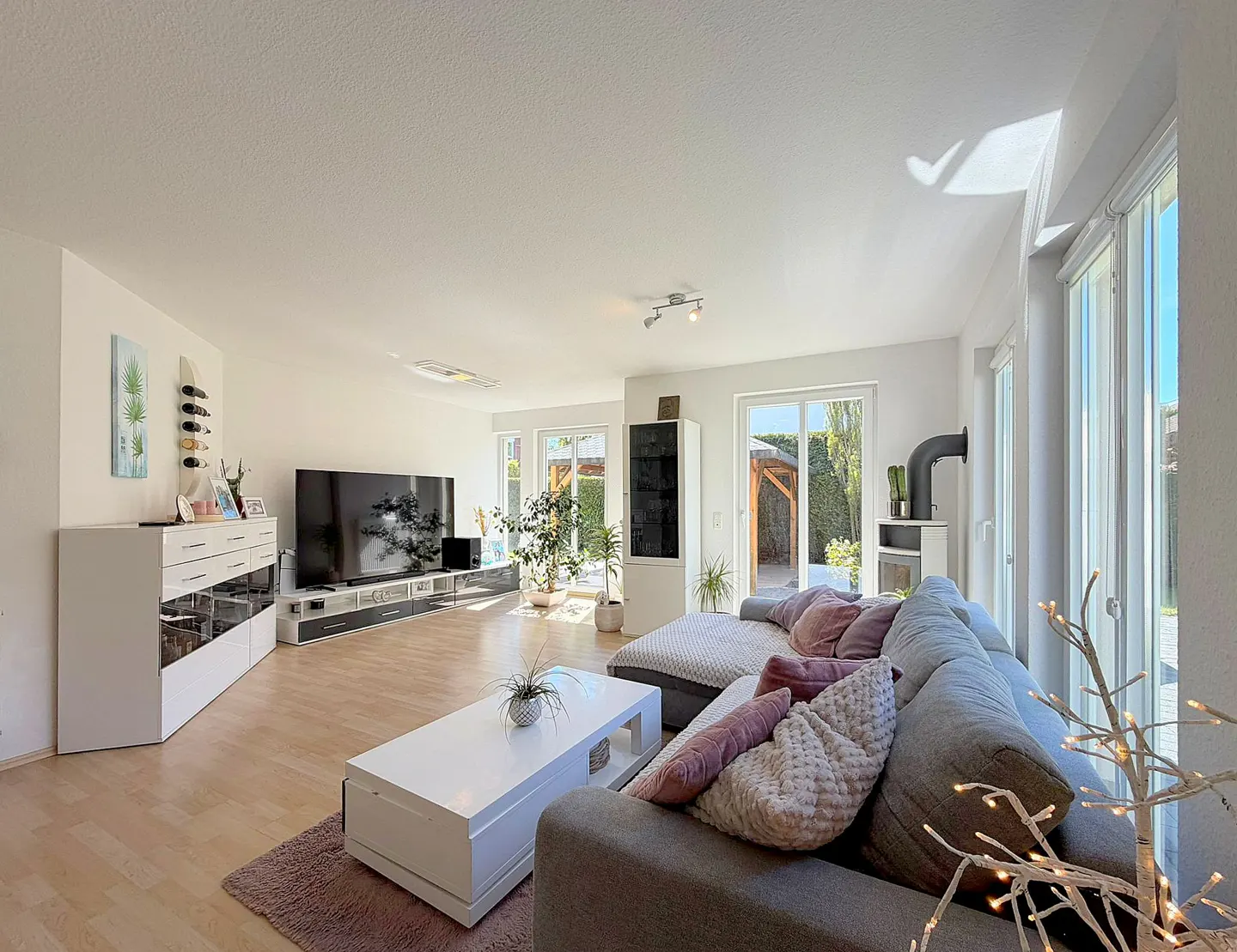 Bright, modern living room with a gray sectional sofa, white coffee table, and large TV. Light wood floors and sliding glass doors to the outside.