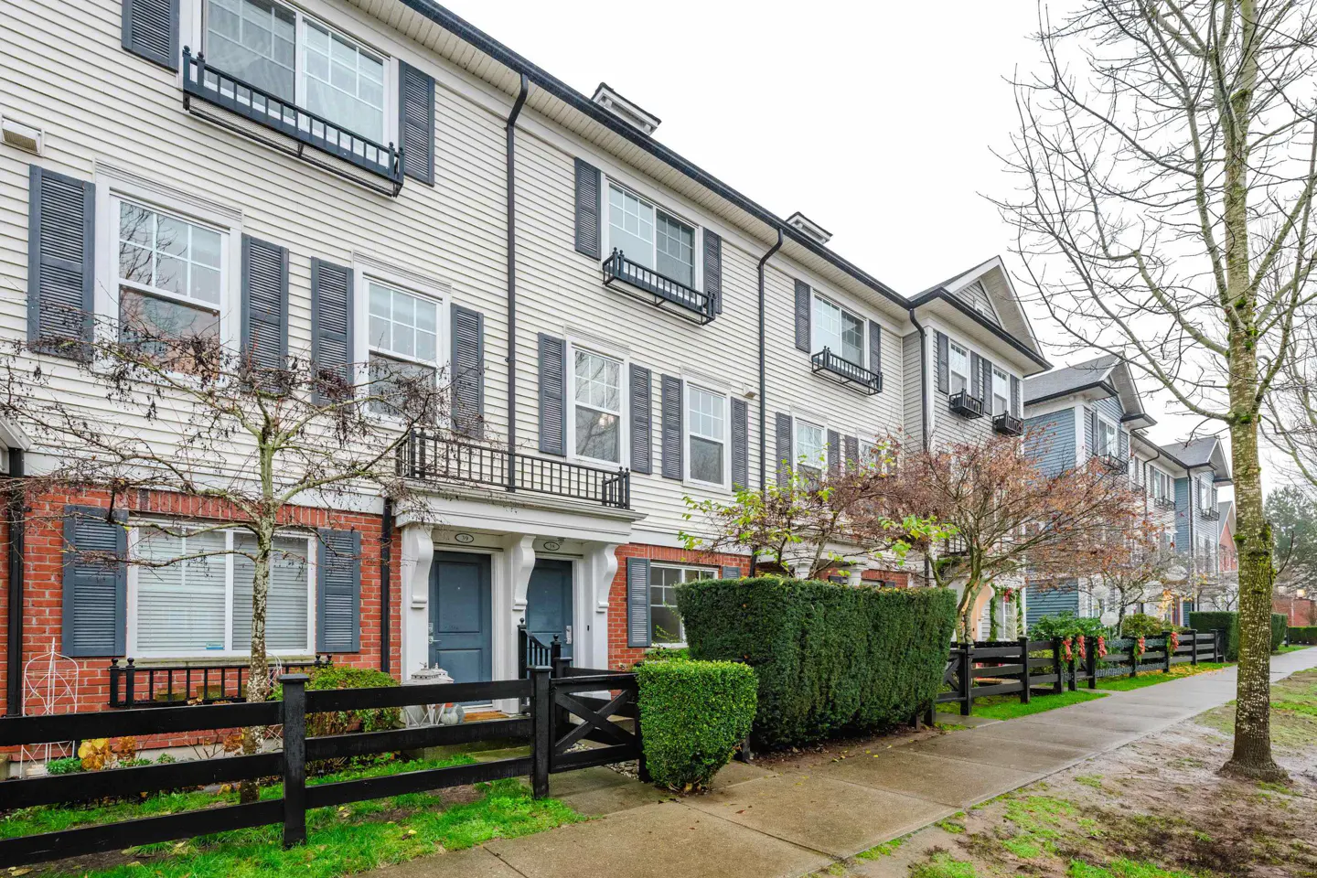 Exterior view of a row of white and brick townhouses with dark shutters, a black fence, and a sidewalk on a cloudy day.