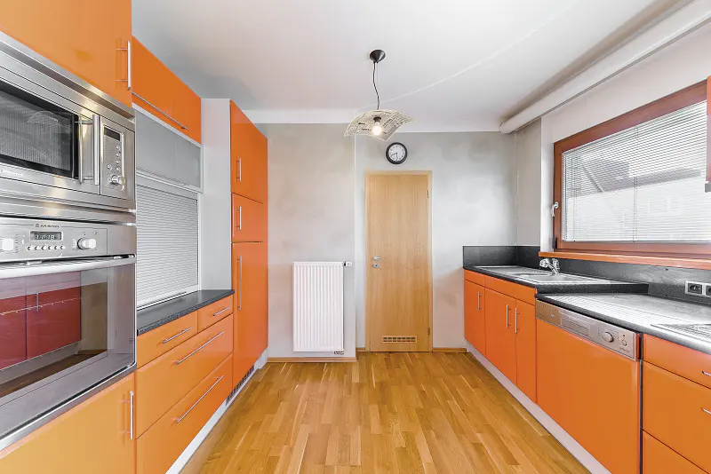 A modern kitchen with orange cabinets, stainless steel appliances, and a light wood floor. A window with blinds is on the right.