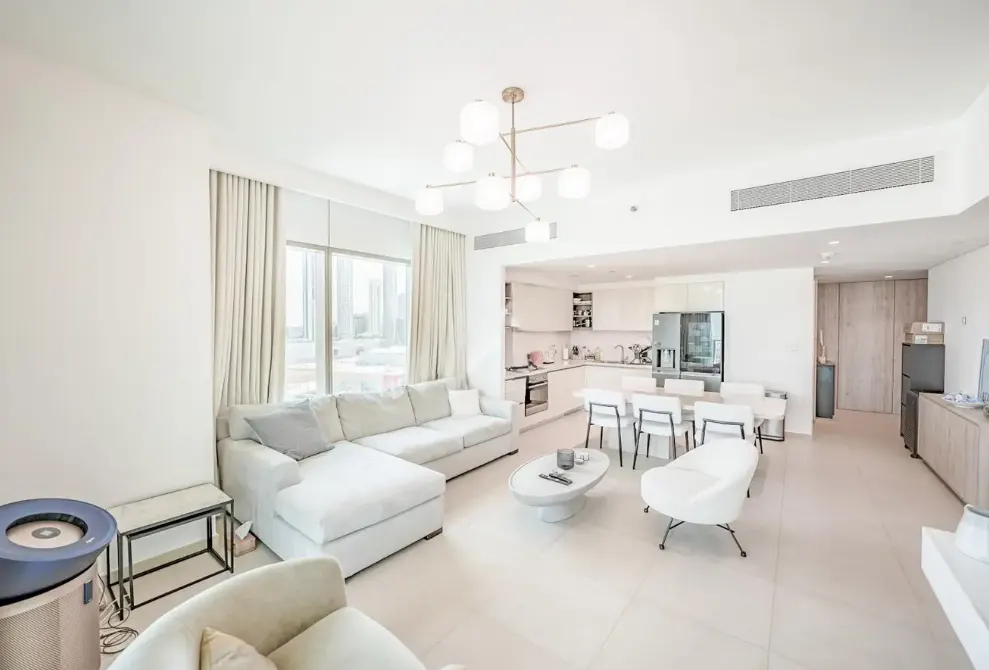 Bright, open-plan living room with a white sectional sofa, modern chandelier, dining area, and sleek white kitchen.