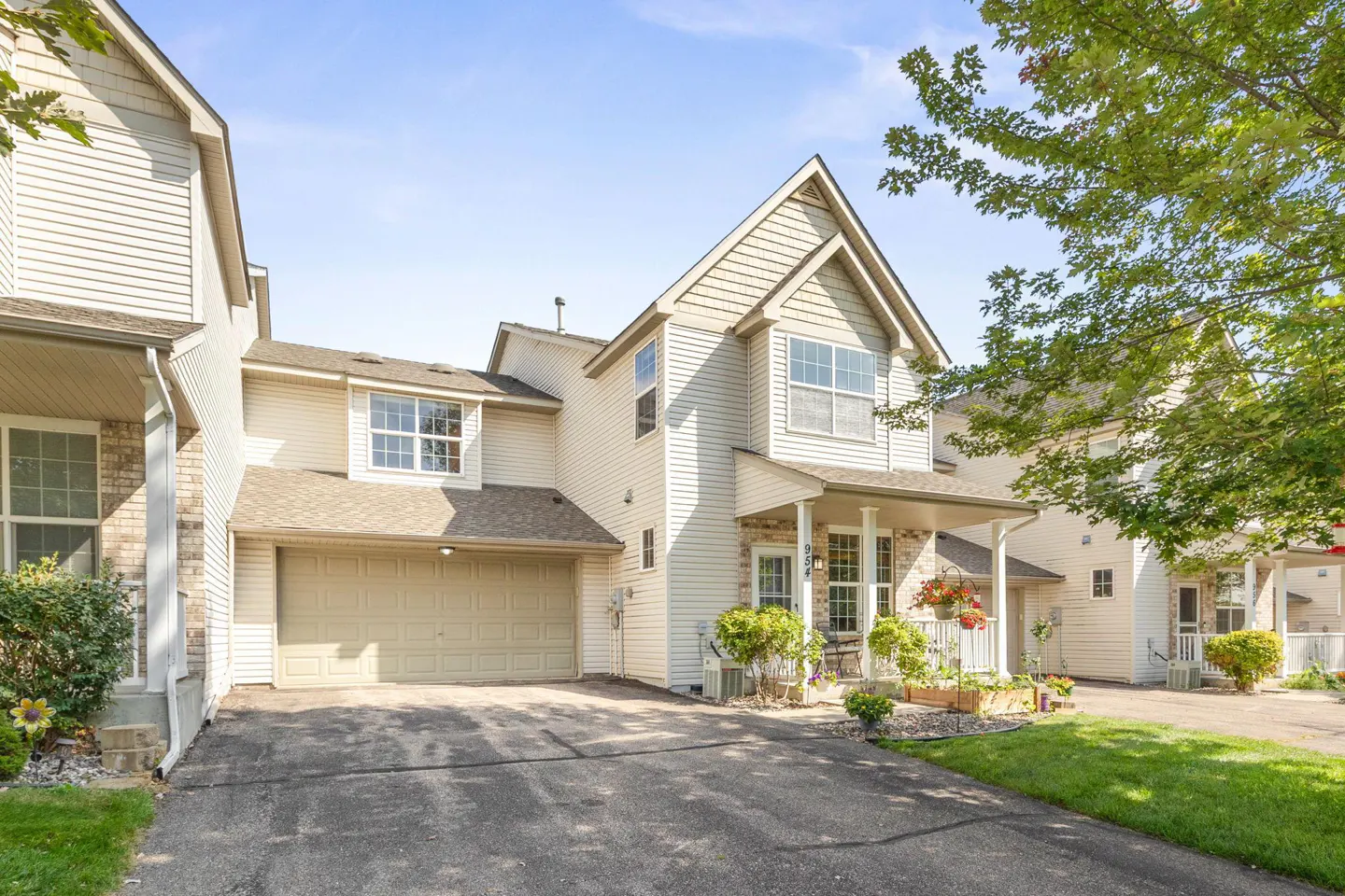 Beige two-story townhouse with a front porch, attached garage, and a driveway. Green trees are visible in the background.