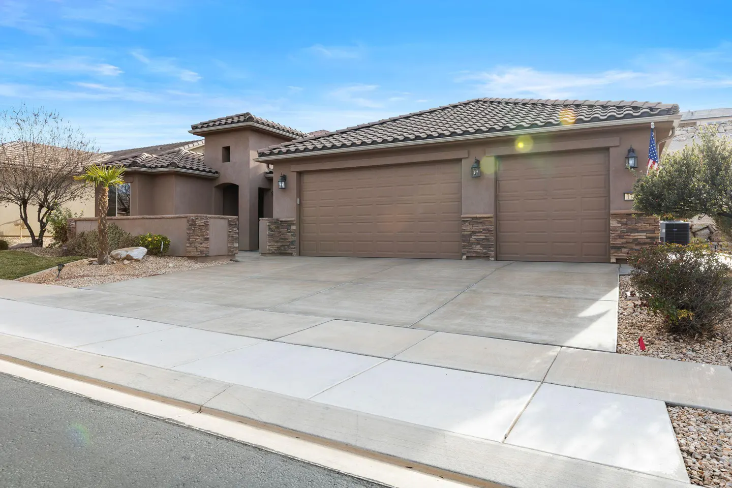 Beige stucco house with a brown tile roof, two-car garage, and a concrete driveway under a blue sky.