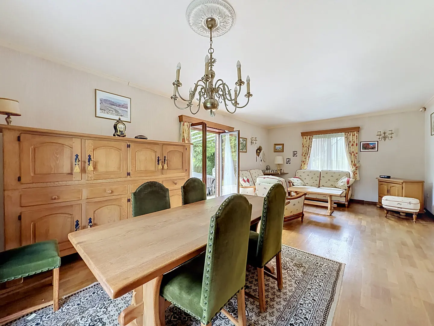 Dining room with wooden table, green velvet chairs, large hutch, chandelier, and a living area with sofa and rug.