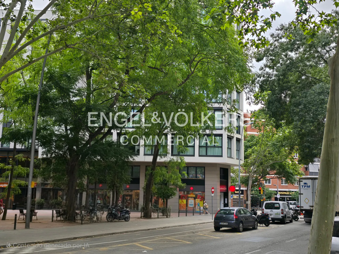 Modern white apartment building with ground-floor shops, surrounded by lush green trees on a city street with parked cars.