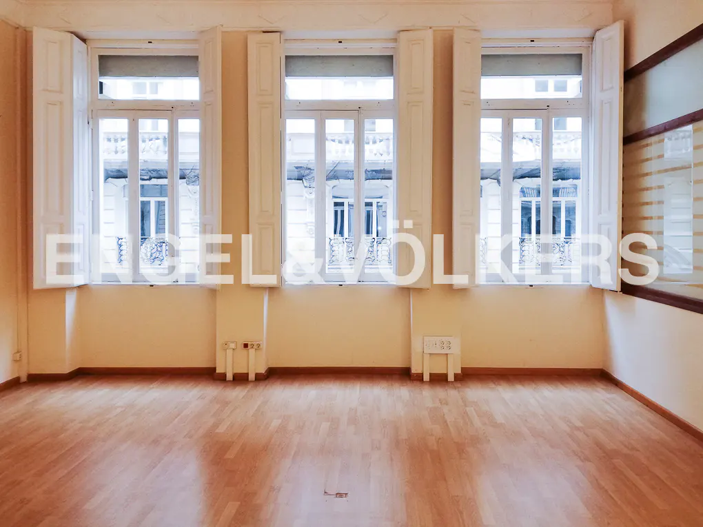 Empty room with light wood floors, cream walls, and three windows with white shutters. The Engel & Völkers logo is superimposed on the image.