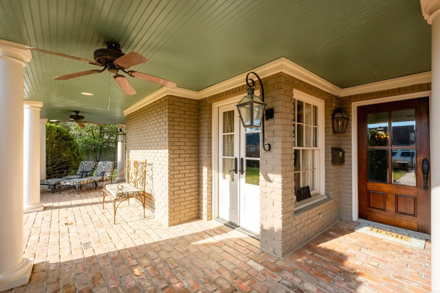 Covered porch with brick floor, white columns, and a green ceiling with a ceiling fan. A white door and a wood door are visible.