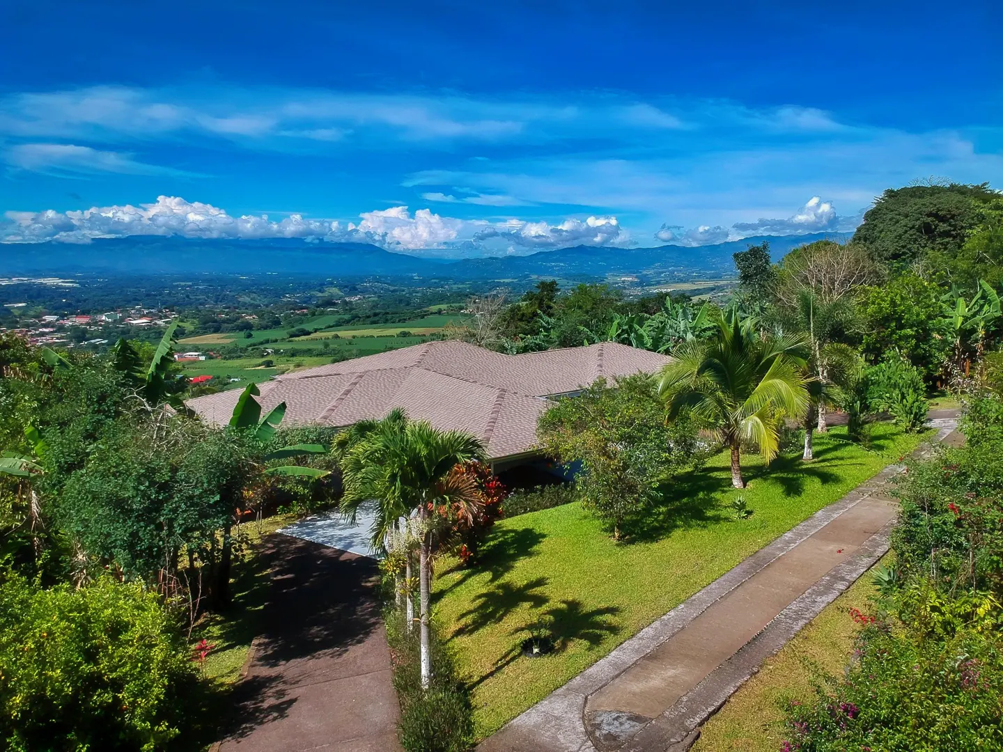 Aerial view of a tan-roofed house surrounded by lush greenery, palm trees, and a green lawn, with mountains and a blue sky in the background.