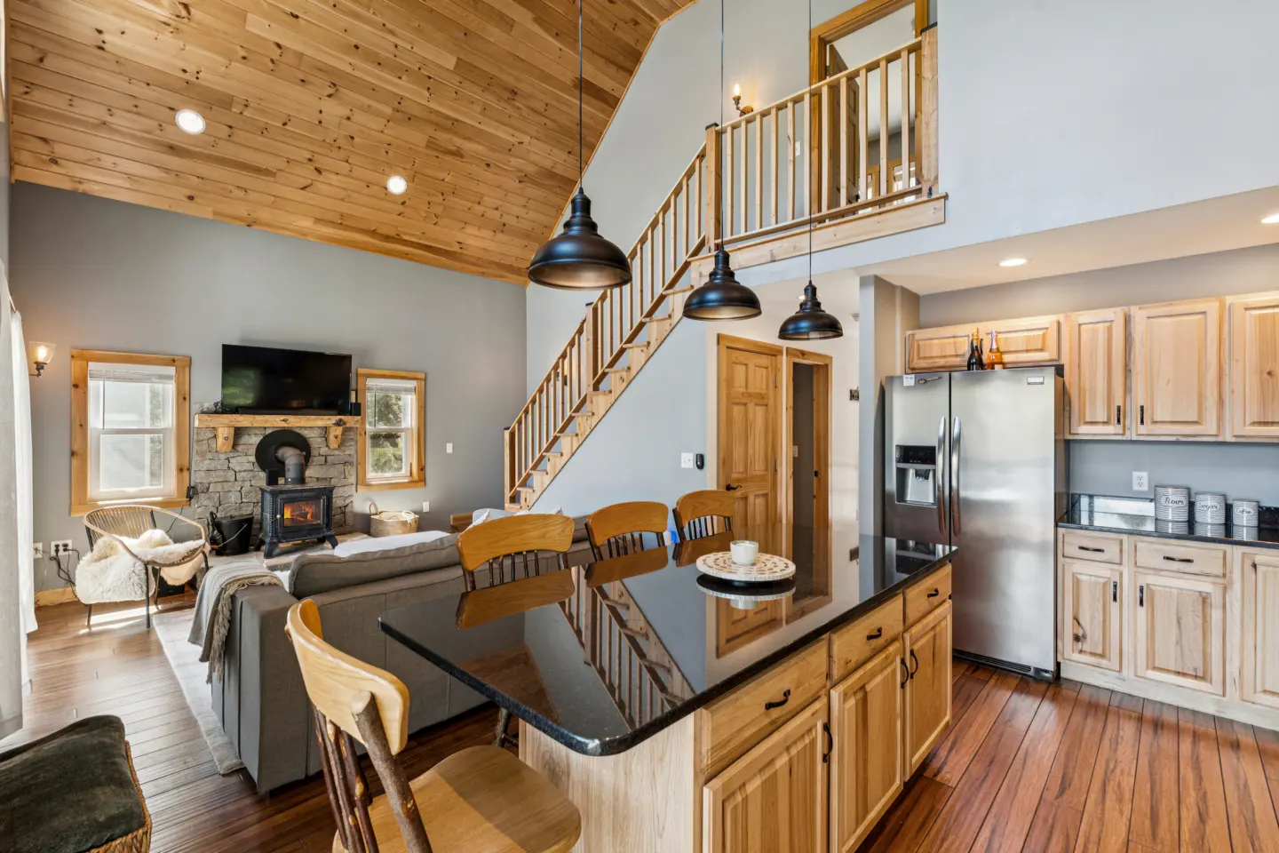Open-concept living space with wood floors, a stone fireplace, and a kitchen island with black countertop. Stairs lead to a loft area.