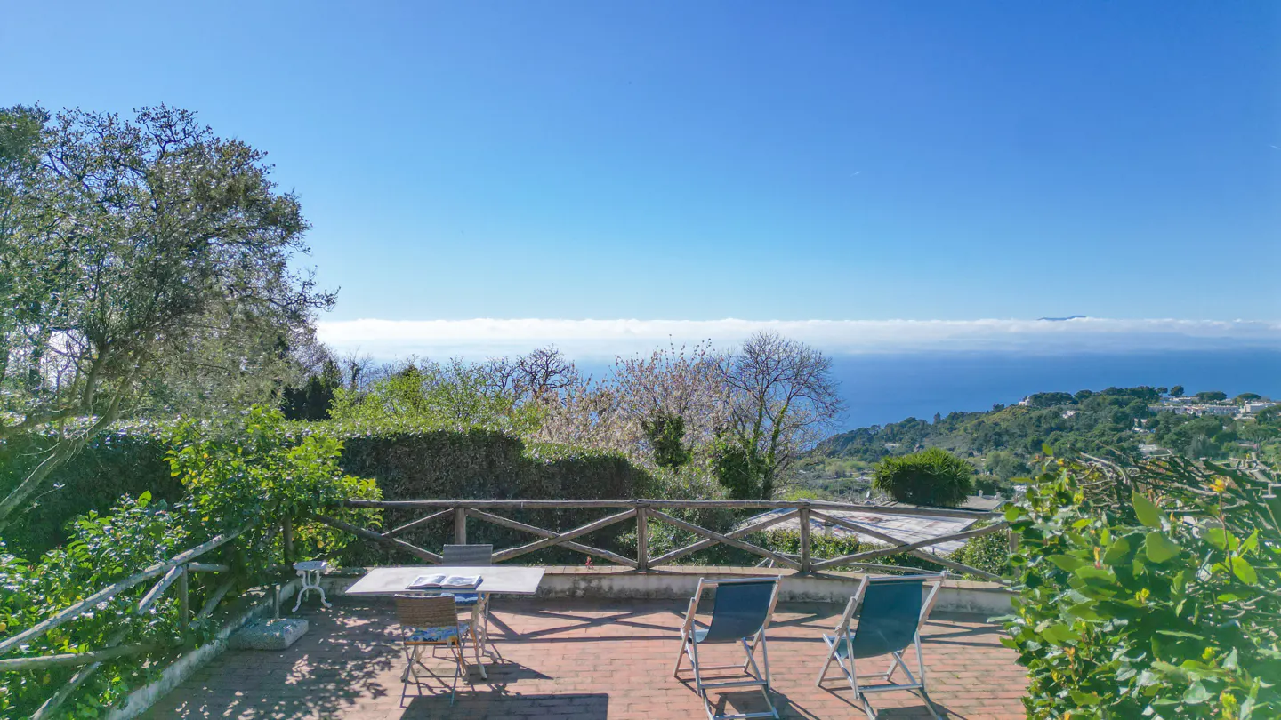 Outdoor patio with table and chairs overlooks a scenic view of the ocean and distant land under a clear blue sky.
